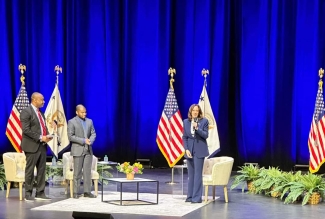 photo of three people on stage, with flags and chairs