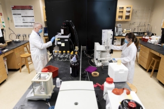 photo of two people on opposite site of lab table, with gloves, coats and masks