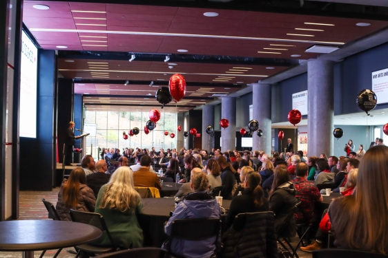 photo of room full of people, balloons, speaker at left