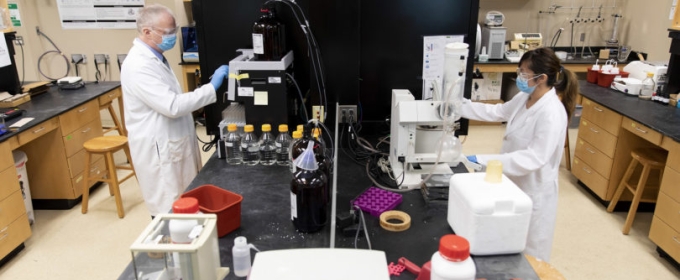 photo of two people on opposite site of lab table, with gloves, coats and masks